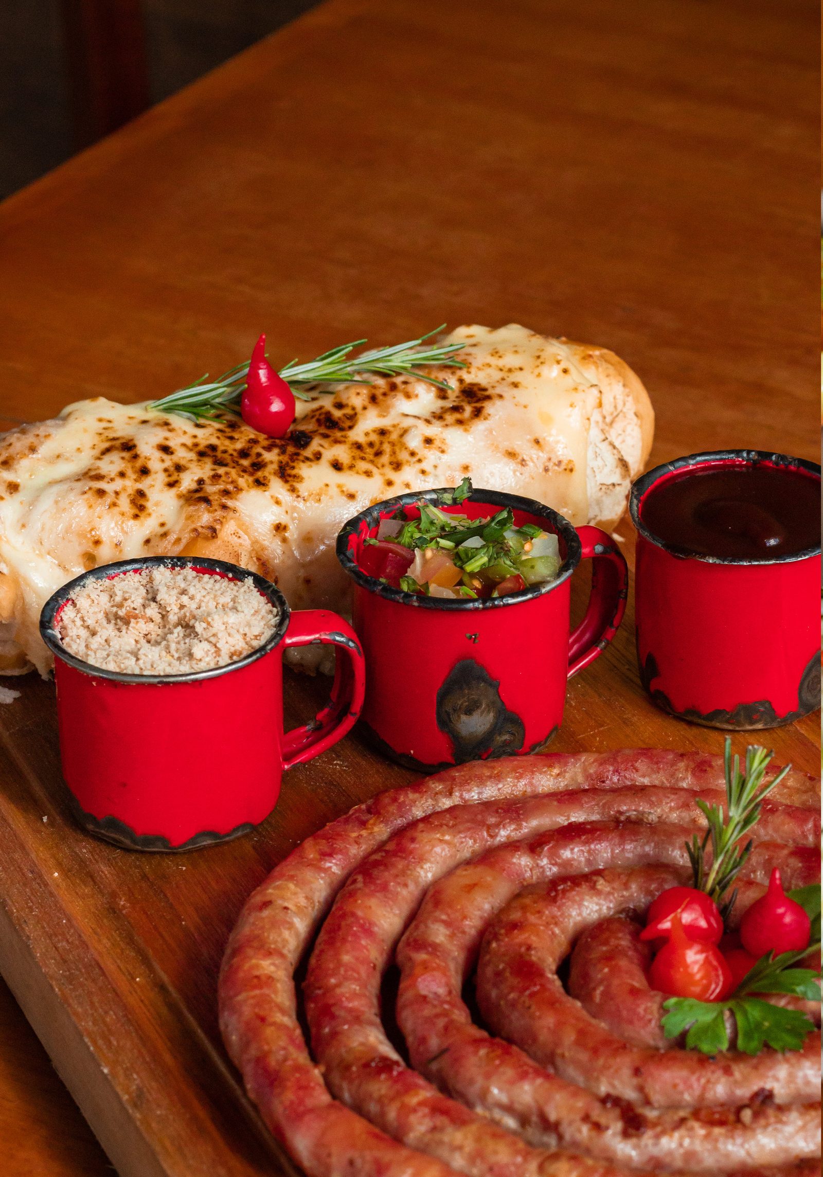 Tábua de linguiça artesanal em espiral sobre madeira escura, com pão de alho gratinado e canecas de vinagrete e barbecue